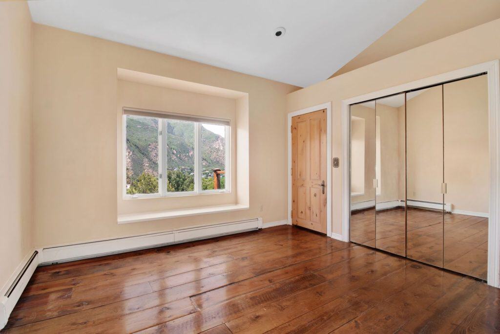 A minimalist, empty room with wooden floors, large window, and mirrored closet reflecting a scenic mountain view.